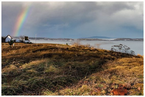 Regenbogen über einer hügeligen Landschaft am Wasser mit vereinzelten Häusern.